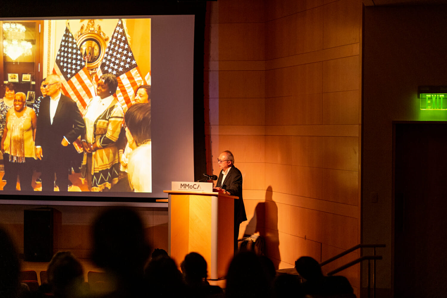 Artist Mel Chin speaking at a podium with a presentation on the projector