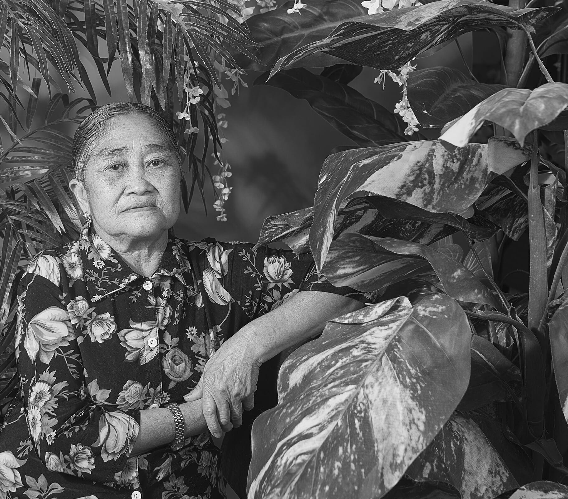 persona mayor con una camisa abotonada con estampado floral, apoyando el brazo contra una superficie, en un entorno frondoso, vegetal y floral