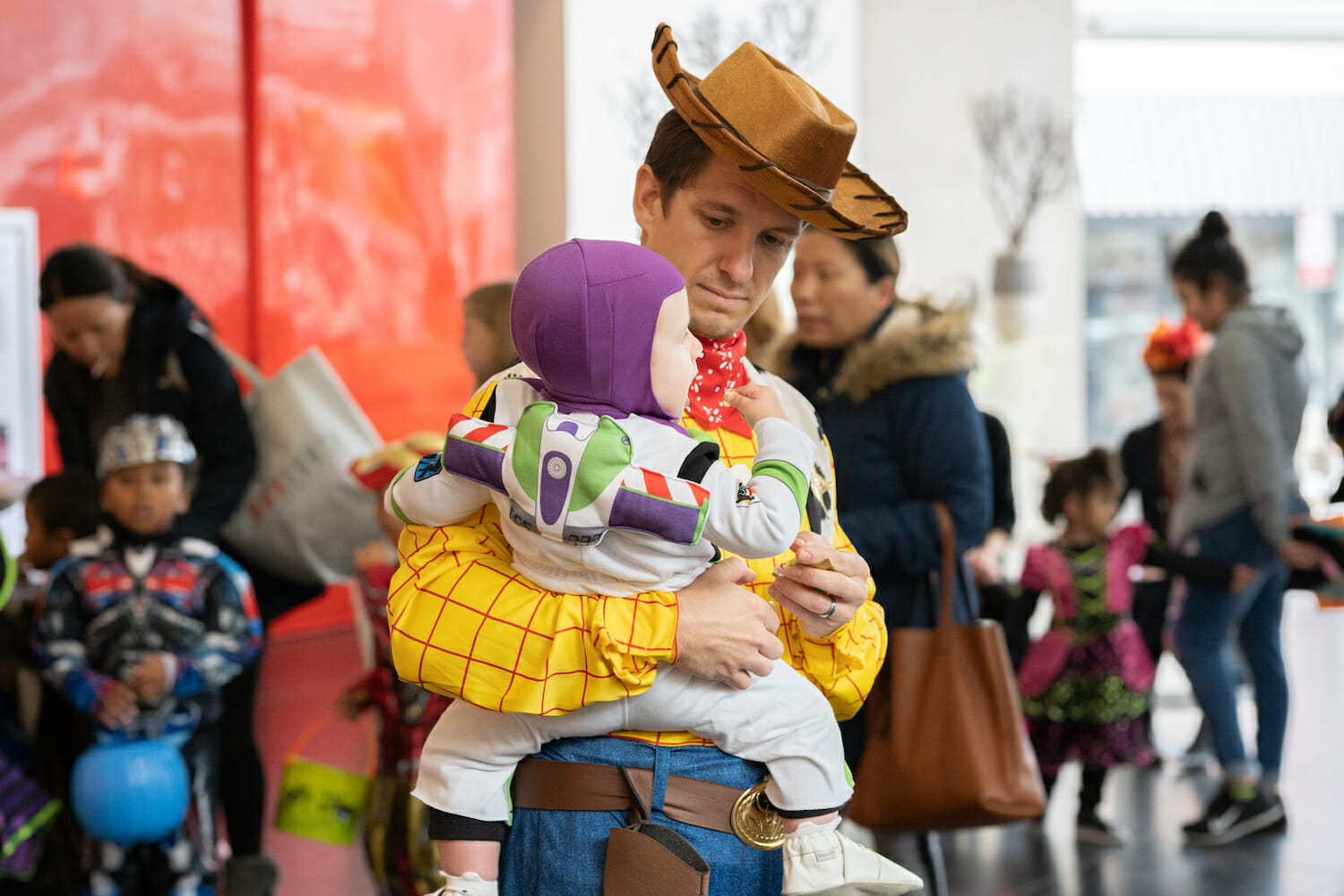 A father holds his son against his hip. Both are wearing costumes from the animated film Toy Story. The father is dressed as Woody and his son is dressed as Buzz Lightyear.