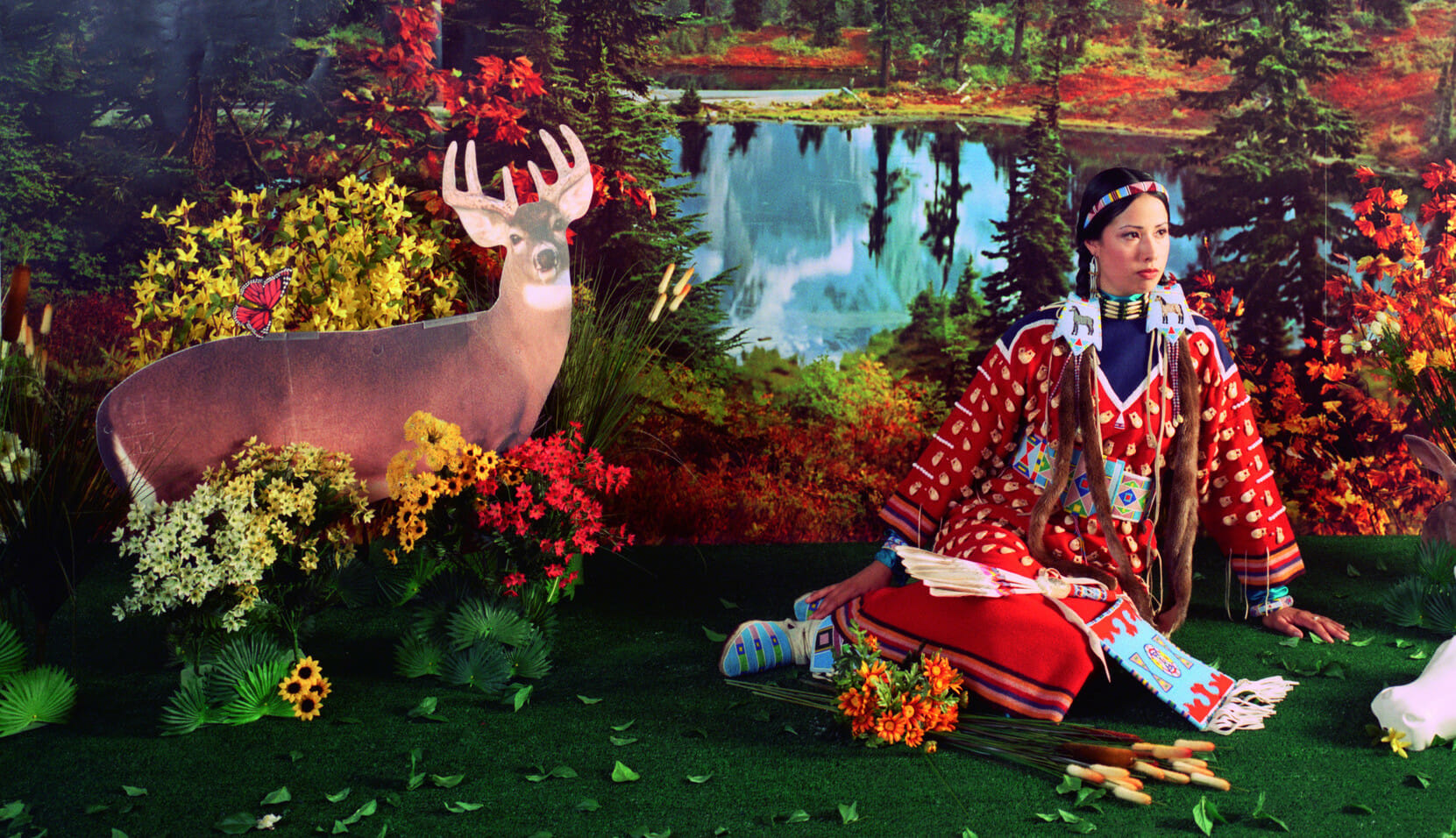 Vestida con ropa indígena tradicional, la artista Wendy Red Star está sentada en un escenario inspirado en un bosque, con recortes de cartón de animales del bosque y plantas de plástico falsas.